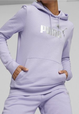 Henkilö pukeutuneena laventelinväriseen Puma-huppariin ja siihen sopiviin collegehousuihin, kädet hupun etutaskussa, yksiväristä taustaa vasten.