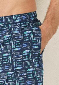 Pantaloni scurți de înot navy cu un talie elastică, având un model de sardine în verde și albastru. Materialul pare a fi ușor și are o textură netedă.