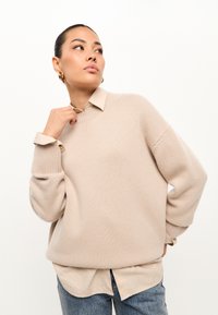 Beige gebreide trui met een ronde halslijn en verlaagde schouders, gedragen over een lichtkleurig button-up shirt, gecombineerd met blauwe jeans.