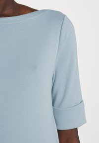 Ljust blå kortärmad topp gjord av mjukt tyg. Har en vid halsringning och uppvikta ärmslut, med en slät yta och enkel design.