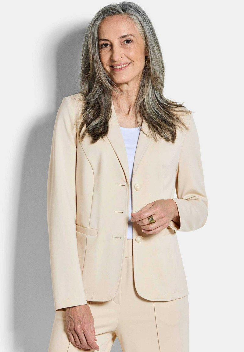 Femme aux longs cheveux gris portant un costume beige et un haut blanc, souriante, avec une bague en or à la main droite.