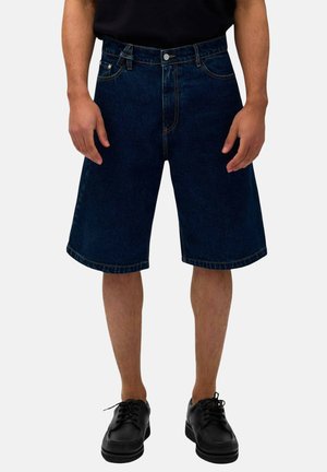 Mand iført mørkeblå denim knælange shorts og sorte snørebåndssko, stående mod en ensfarvet baggrund.
