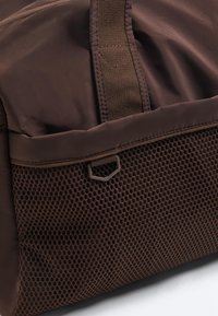 Zbliżenie na brązowy plecak z tkaniny z siatkową kieszenią i wzmocnionym paskiem przeszytym pionowo nad kieszenią.