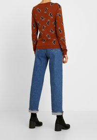 Un pull marron présentant un motif noir de chevaux répété, associé à un jean en denim bleu clair. Le jean est à coupe droite avec des poignets retroussés.