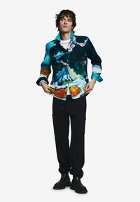 Košeľa na zapínanie s viacfarebnými ovocnými a kvetinovými motívmi, vyrobená z látky. Čierne nohavice a členkové čižmy, modelka pózuje na bielom pozadí.