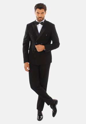 Tuxedo nero doppiopetto con rever in raso, camicia bianca e cravatta a farfalla. Scarpe lucide nere completano il look formale.