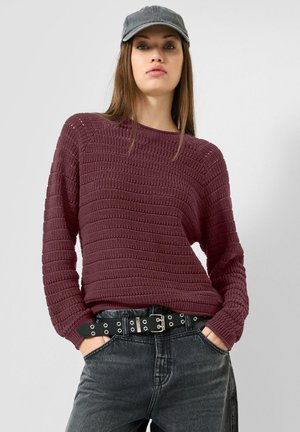 Jeune femme portant une casquette grise, un pull en maille bordeaux, une ceinture noire avec des œillets argentés et un jean gris foncé, les mains dans les poches.