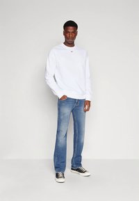 Hombre vestido con sudadera blanca, jeans azules y zapatillas negras, de pie contra un fondo claro y liso con una mano en el bolsillo.