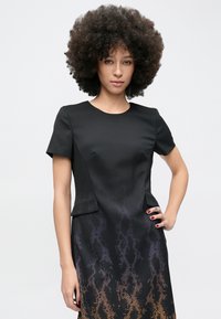 Femeie cu păr creț purtând o rochie neagră mulată cu mâneci scurte și un model degradat de piele de șarpe, stând cu o mână pe șold.