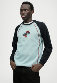 Ανοιχτό μπλε φούτερ με μαύρα μανίκια, κόκκινες ραφές και κεντητό λογότυπο. Διαθέτει μανίκια ράγκλαν και στρογγυλή λαιμόκοψη.