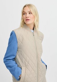 Beige gesteppte Weste mit hohem Kragen, Reißverschluss vorne und seitlichen Taschen. Blaues langärmliges Hemd unter der Weste, kontrastierende Farben und Texturen.