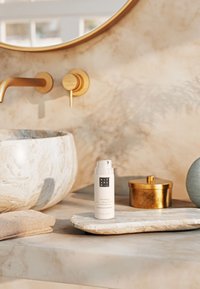 Bouteille cylindrique blanche de shampooing sec sur un plateau en pierre claire texturée, à côté d'un conteneur rond doré et d'une pierre grise, avec un évier en marbre en arrière-plan.