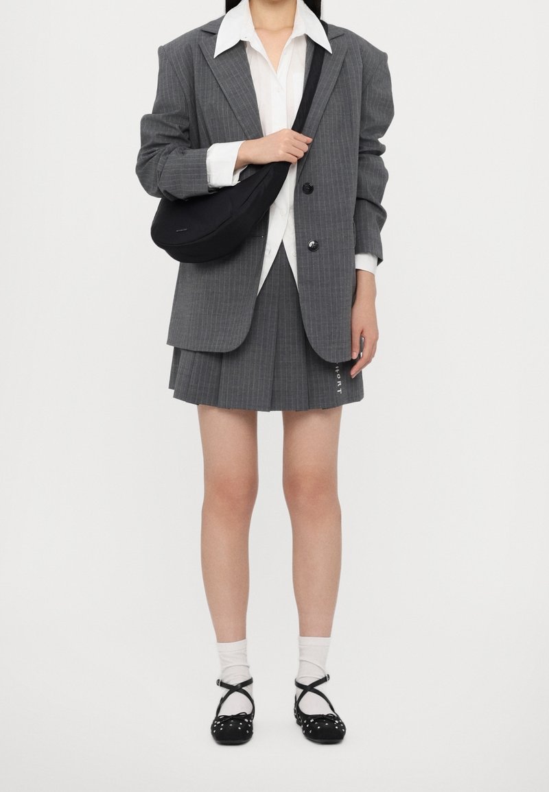 Person iført grå pinstripe blazer og plisseret nederdel, hvid skjorte, bærende sort crossbody taske, med hvide sokker og sorte ballerinasko.