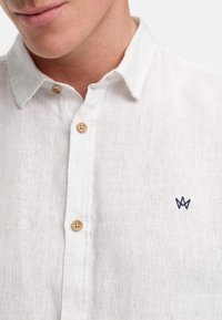 Wit linnen shirt met een button-down kraag, houten knopen en een klein marineblauw geborduurd logo op de borst. Zachte textuur, lichtgewicht stof.