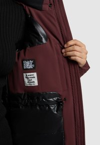 Chaqueta exterior burdeos con forro interior negro, dos etiquetas y un bolsillo con cierre de cordón para mayor funcionalidad.