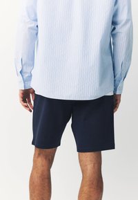 Jasnoniebieska koszula w paski z długim rękawem i zapinanymi mankietami, noszona z granatowymi szortami. Tkanina wydaje się gładka, z casualowym fasonem i linią wykończenia.