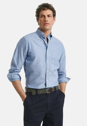 Camisa de botones azul claro con mangas enrolladas y un bolsillo en el pecho, combinada con pantalones azul marino y un cinturón verde con una hebilla de plata.
