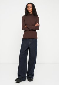 Vrouw met krullend haar staat met gekruiste armen, gekleed in een bruine coltrui, wijdvallende donkere jeans en zwarte plateau-schoenen, tegen een witte achtergrond.