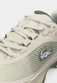 Κοντινό πλάνο ανοιχτόχρωμου πράσινου αθλητικού παπουτσιού Lacoste με υφή από δίχτυ, λευκά κορδόνια και κεντημένο λογότυπο κροκοδείλου στο πλάι.