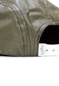 Grünes Stoffmützchen mit glatter Textur, kontrastierenden schwarzen Nähten, einem weißen Riemen und einem Markenetikett an der Seite.