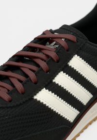 Black athletic shoe featuring maroon laces, white serrated stripes, textured upper, and a textured sole with a brown accent.
