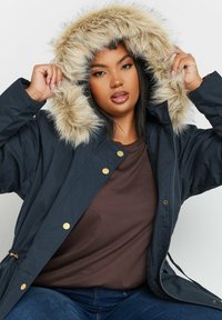 Mörkblå parka med fuskpäls på huvan, guldiga detaljer och ett kviltat mönster. Har dragkedja och justerbar midja.