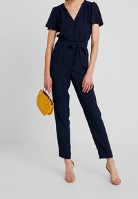 Mujer vestida con un mono azul marino de manga corta y cinturón anudado en la cintura, sosteniendo un bolso color mostaza y sandalias de tacón nude.