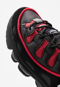 Baskets de sport noires et rouges avec une tige en cuir texturé, une languette rembourrée, un design de lacets unique et un amorti à air visible dans la semelle.
