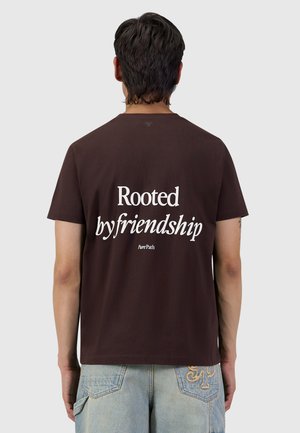 Man met donker haar die een donkerbruin T-shirt draagt met de tekst "Rooted by friendship" en lichtblauwe spijkerbroek met borduurwerk op de achterzakken.