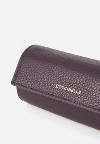 Lila strukturiertes Lederetui mit abgerundeten Kanten und einem goldenen "Coccinelle"-Logo. Glattes Innenleben, designed für das Halten von Brillen oder kleinen Gegenständen.