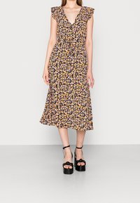 Femme portant une robe midi noire à fleurs avec des manches volantes et une taille à nouer, associée à des sandales à plateforme noires avec des brides de cheville.