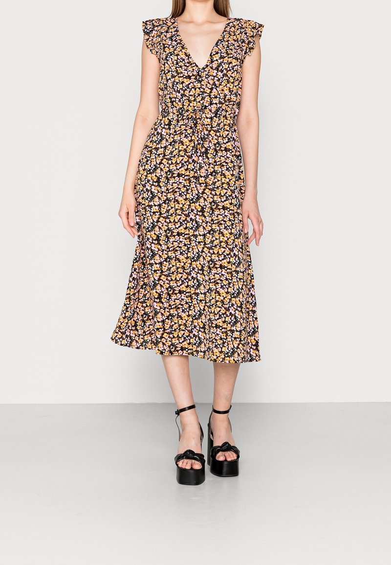 Femme portant une robe midi noire à fleurs avec des manches volantes et une taille à nouer, associée à des sandales à plateforme noires avec des brides de cheville.