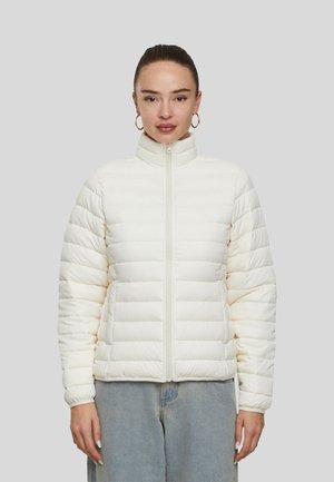 Femme portant une veste matelassée blanche à col montant et un jean bleu, debout devant un fond uni gris clair.