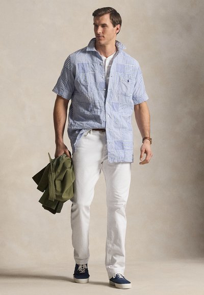 Άνδρας με ανοιχτό μπλε πουκάμισο με σχέδια και κοντά μανίκια, άσπρο παντελόνι, ναυτικά αθλητικά παπούτσια, κρατώντας διπλωμένο χακί μπουφάν, στέκεται μπροστά σε ουδέτερο φόντο.