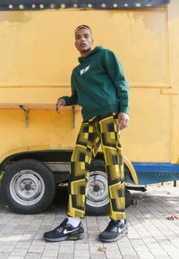 Groene hoodie met logo, gemusterde gele en zwarte broek, zwarte sneakers met groene accenten, staand naast een gele trailer.