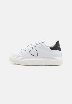 Vit lädersneaker med rund tå, tjock vit sål, svart hälsaccent och en logotypflik på sidan. Snören framtill.