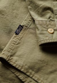 Camisa verde oliva con una textura suave, que presenta un cierre de botón y costuras visibles. Una etiqueta oscura con "Superdry" está cosida en la tela.