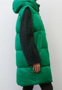 Grøn puffy jakke med hætte, der har mørke strikkede ærmer, lynlåsfortil og quiltet tekstur. Fremhævet af en livlig farve.