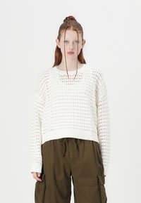 White, knitted sweater with a loose design, featuring a textured pattern, ribbed cuffs, and a cropped hem, paired with olive cargo pants.