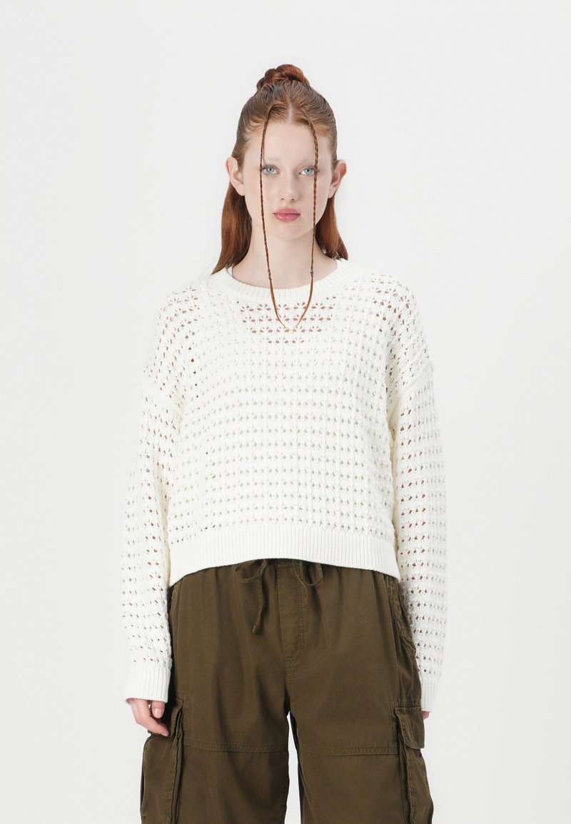 White, knitted sweater with a loose design, featuring a textured pattern, ribbed cuffs, and a cropped hem, paired with olive cargo pants.