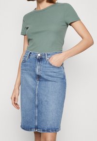 Top de manga corta verde acanalado combinado con una falda de mezclilla azul hasta la rodilla. La falda cuenta con un pliegue en el frente, bolsillos laterales y costuras visibles.