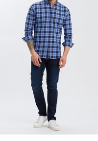 Muž v modro-černě kostkované košili s dlouhým rukávem, tmavě modrých džínách a bílých teniskách, který ukazuje tetování na předloktí na bílém pozadí.