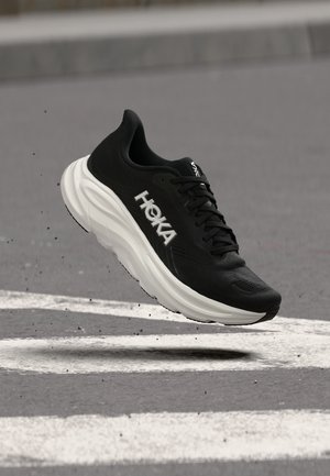 Scarpa da corsa nera Hoka con suola bianca che fluttua sopra una strada lastricata con segnaletica bianca.