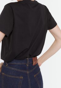 Sort t-shirt med korte ærmer, der er parret med højtaljede blå denimjeans, som har et lædermærke på den bagerste talje og gule sømme.