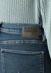 Džinsu bikses ar tumši zilu mazgāšanu, ar G-Star Originals ādas uzlīmi jostasvietā un teksturētu gaiši zilu adījumu augšdaļu.