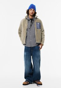 Mlad oseba, oblečena v modro pleteno kapo, bež flis jakno z žepom na prsih, črno-belo karirano srajco, široke modre kavbojke in svetlo rjave pohodne čevlje.