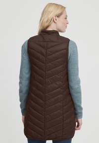 Brun quiltet vest med høj krave, der har et chevron-mønster og letvægtsmateriale. Bæres over en blå strikket sweater.