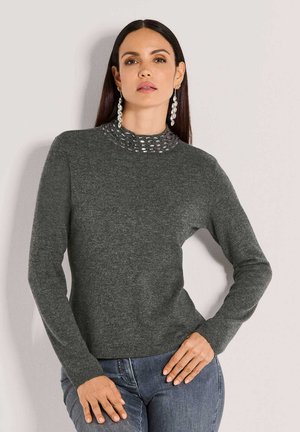 Frau mit langen dunklen Haaren, die einen grauen Strickpullover mit metallischem Kragen und eine blaue Jeans trägt, posiert vor einer hellen Wand.
