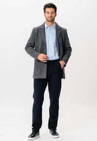 Mand står iført en grå blazer, lyseblå stribet skjorte, mørke bukser og sorte sneakers med hvide såler foran en hvid baggrund.