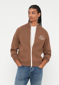 Brun zip-up hettegenser med hvit glidelås, ribbestrikkede mansjetter, og "REPLAY MOTORCYCLE COMPANY" trykket på brystet. Casual passform med sidelommer.